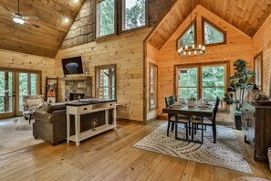 A log cabin interior with a fireplace, dining area, and large windows.