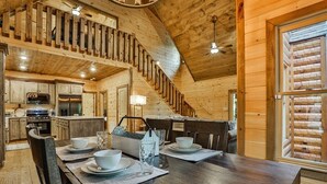 A wooden interior with a dining table set for four, kitchen area with appliances, and a staircase. - Happy Hideaway | Mineral Bluff, Ga (Mineral Bluff)