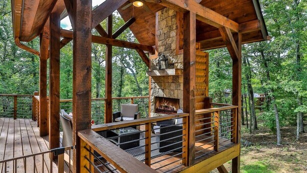 A wooden deck with a stone fireplace, outdoor seating, and a view of the surrounding forest. - Happy Hideaway | Mineral Bluff, Ga (Mineral Bluff)