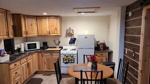 Cocina compacta con armarios de madera, un microondas, estufa y refrigerador. Hay una mesa de comedor con sillas y un florero con flores.