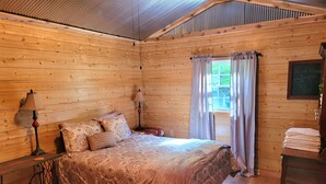 Una habitación de cabaña de madera con una cama, dos lámparas de noche, un ventanal con cortinas y una pizarra con un mensaje.