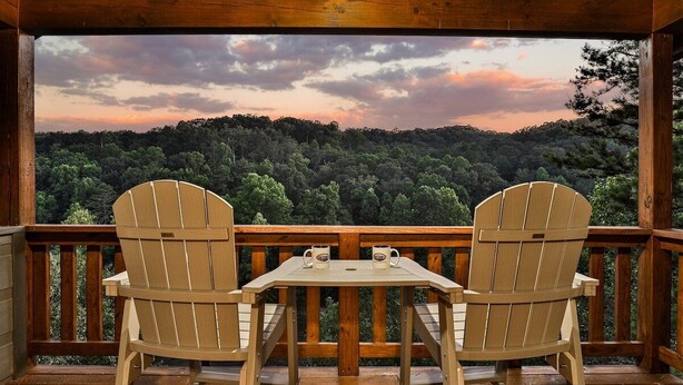Eine Holzdiele mit zwei Adirondack-Stühlen und einem Tisch, mit Blick auf eine bewaldete Landschaft.