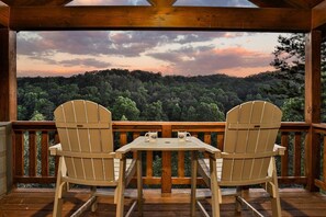 Een houten terras met twee Adirondack-stoelen en een tafel, met uitzicht op een bebost landschap.
