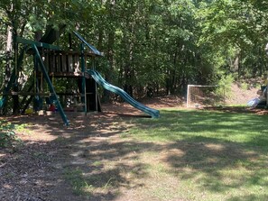Explore a natural playground surrounded by trees, featuring a play structure with a slide and swing set on a grassy lawn.