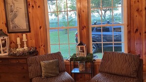 A cozy room with wooden walls and floors, featuring two striped armchairs, a chandelier, and a large window with a view of the outdoors. - Creekside Country Cottage- minutes away from I-95 (Manning)