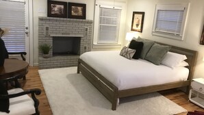 Een slaapkamer met een bakstenen open haard, een bed met veel kussens, een nachtkastje en een raam met jaloezieën.