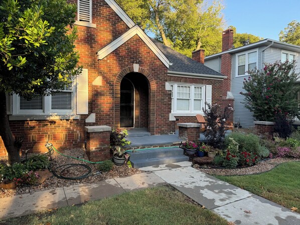 A brick house with a front porch, a garage door, and a small garden with various plants and flowers. - Best Location, comfy bungalow just a 10 min.walk to OU Stadium or Campus Corner (Norman)