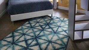 Ein Schlafzimmer mit Bett, einem Teppich mit geometrischem Muster, einem Fenster mit Vorhängen und einem Bücherregal.