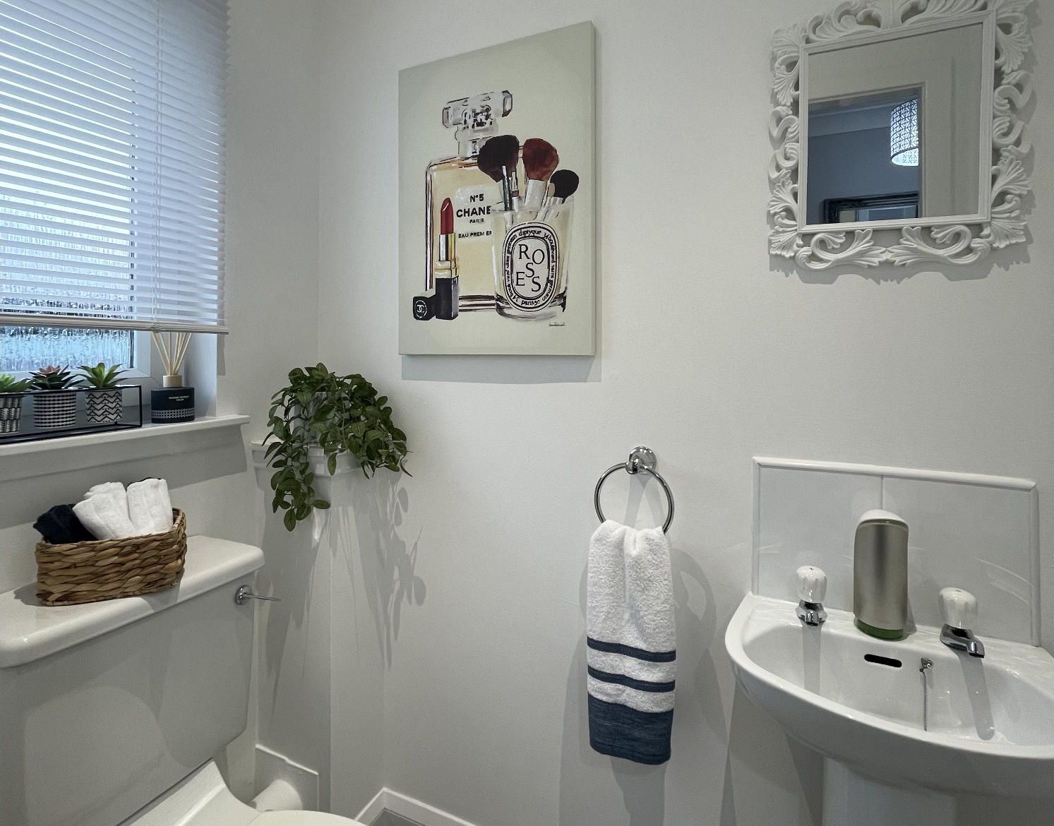 A bathroom with a toilet, sink, and window with blinds. There is a towel rack with a towel, a potted plant, and a decorative mirror.