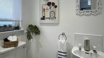 A bathroom with a toilet, sink, and window with blinds. There is a towel rack with a towel, a potted plant, and a decorative mirror.