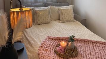 Un dormitorio ordenado con cama, almohadas, una lámpara de noche, un florero con plumas y una bandeja con velas y una planta.