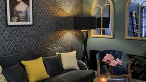 A room with a patterned wall, a dark sofa with yellow cushions, a black floor lamp, and a blue armchair with a vase of pink flowers. - 2 Bed House - Sleeps 4 - Garden, Parking, Wifi (Perth)