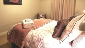 A bedroom with a bed, pillows, a folded towel, a chair, a wall art piece, and a window with curtains. - Your Home Away From Home in a Charming Street (West Launceston)