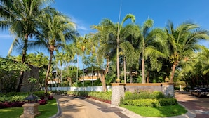 Entrada de um resort tropical, com palmeiras, um caminho de pedra e um jardim bem cuidado.