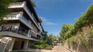 Um edifício residencial com vários andares, varandas, entrada pavimentada e vegetação ao redor.