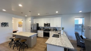 A modern kitchen with white cabinets, stainless steel appliances, and a marble countertop. There are three stools at the island and a dining area with a chair. - New home-3 miles to DT Flag!! With grill, firepit, AC (Flagstaff)