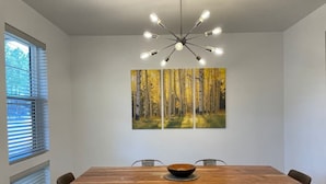 A dining area with a wooden table, four chairs, a central light fixture, and two paintings on the wall. - New home-3 miles to DT Flag!! With grill, firepit, AC (Flagstaff)
