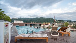 A rooftop pool area with wooden loungers and a view of a cityscape and hills in the background. - Ozone Condo by Lofty (Karon)