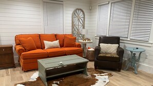 Ett rum med trägolv, en brun och vit matta av kohud, en orange soffa, en svart fåtölj, ett litet runt bord och en dekorativ träskulptur på väggen.