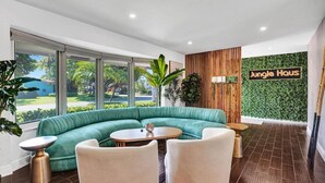 Un espace de détente moderne avec un canapé turquoise aux lignes courbes, deux fauteuils blancs, une table basse ronde en bois et une petite table d’appoint. Un panneau indiquant « Jungle Haus » est visible sur le mur.
