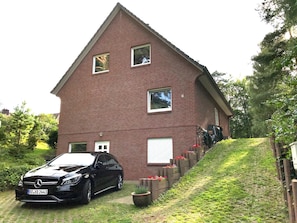 A brick house with a car parked in front, a small garden with flowers, and a sloped driveway. - Relaxation in the Forest Apartment Hamburg 1 (Rosengarten)