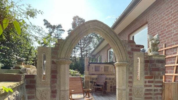 Un arco de piedra que da paso a una terraza de madera con una mesa y sillas, rodeada por un muro de ladrillo y vegetación.