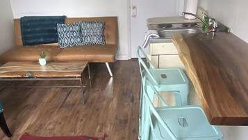 A modern living room with a brown sofa, a small table with a plant, and a colorful rug. Adjacent is a kitchen with a white stove, sink, and light blue bar stools.