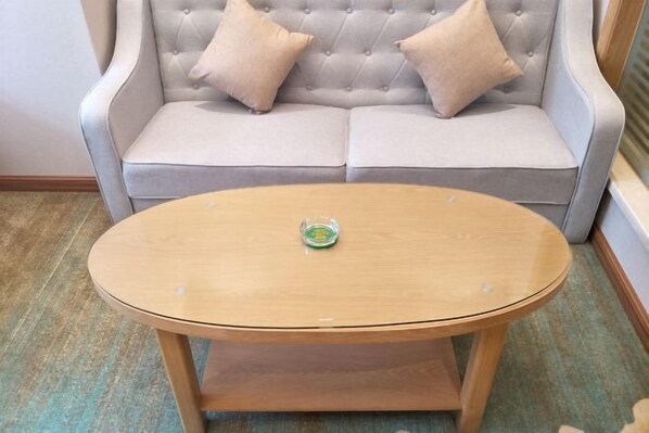A round wooden table with a glass object on it, placed in front of a grey tufted sofa with two beige cushions. - Vienna Hotel (Qingyuan Zhongshan Park Century Plaza) (Qingyuan)