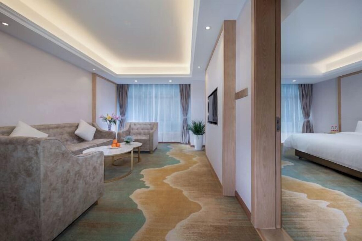 A modern hotel room with a sofa, a small table with a vase, a bed, and a large rug.