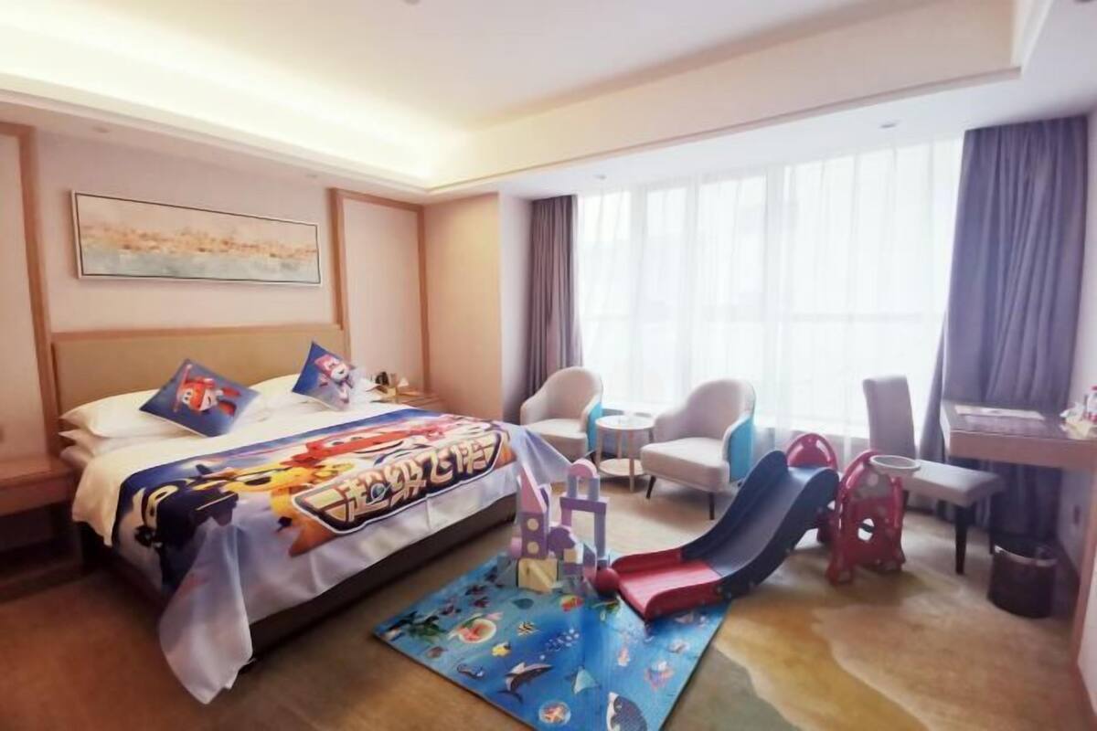A hotel room with a bed, a desk, a chair, a small table, a window with curtains, and a play area with a slide and a mat.