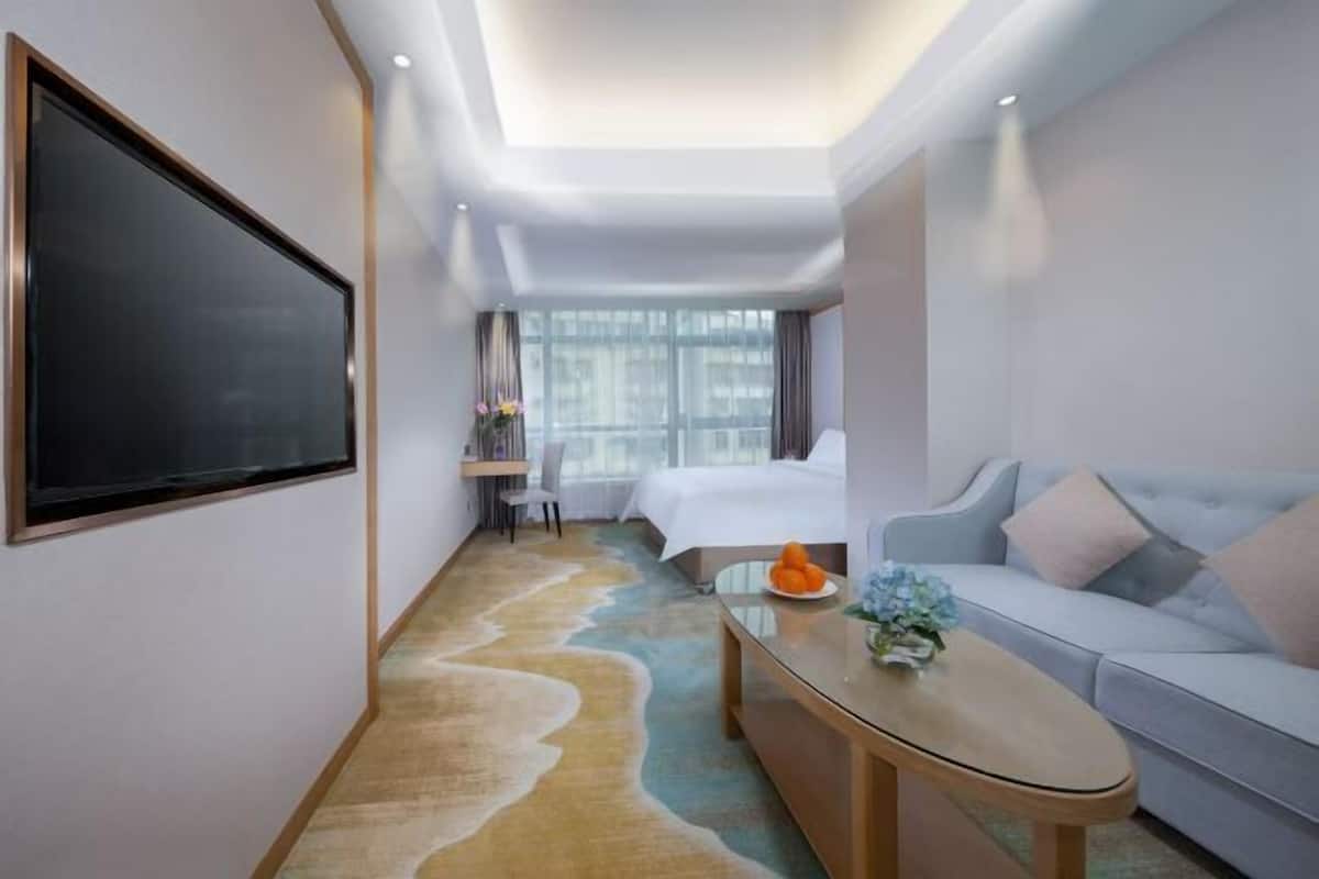A hotel room with a large flat-screen TV, a bed, a sofa, a desk with a chair, and a window with curtains.