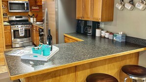 A kitchen with wooden cabinets, stainless steel appliances, a granite countertop, and bar stools. - Cozy Cabin Themed Basement (Morgan)