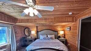 A wooden-paneled bedroom with a bed, two bedside tables, a ceiling fan, and a window with curtains. - Cozy Cabin Themed Basement (Morgan)