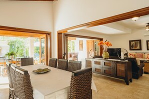 Un comedor amplio con una mesa grande y sillas de mimbre, una zona de cocina con televisor y objetos decorativos, y vistas al exterior a través de amplios ventanales.