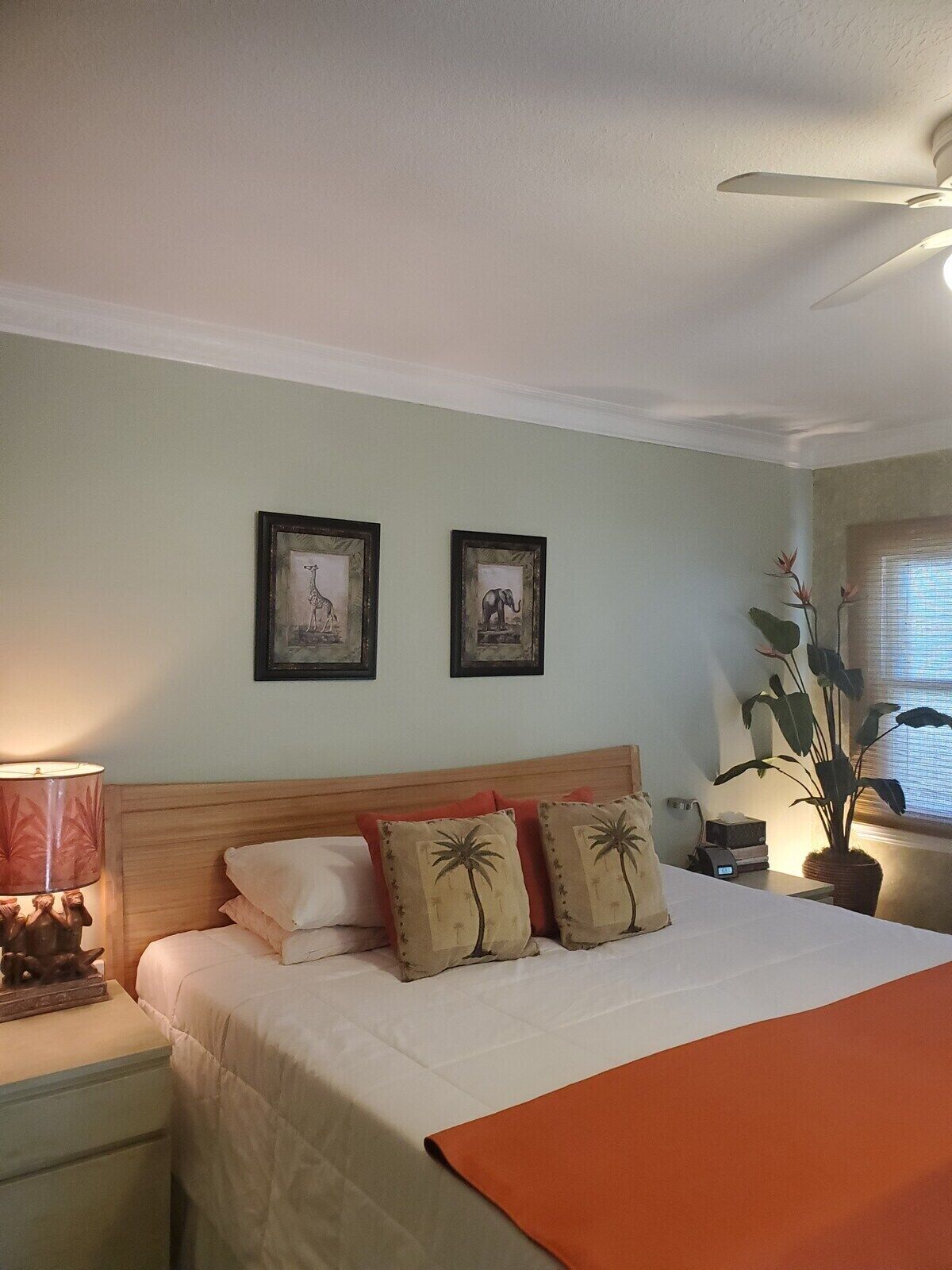 A bedroom with a bed, pillows, a nightstand, a lamp, a ceiling fan, and framed pictures on the wall.