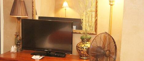 A wooden dresser with a television, a mirror, a vase with branches, and a standing fan.