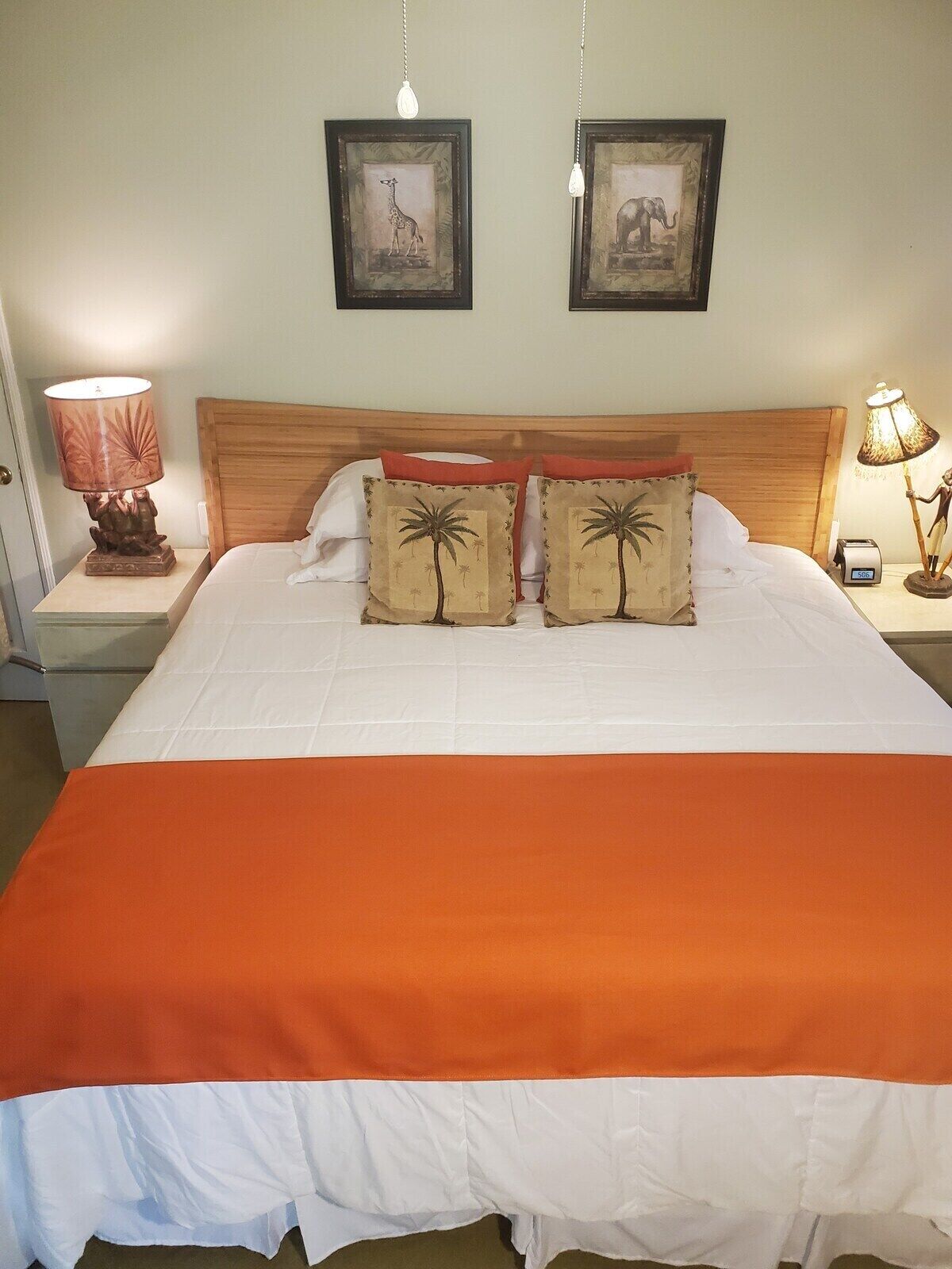 A neatly made bed with an orange bedspread, two matching pillows with palm tree designs, and two framed pictures on the wall above.