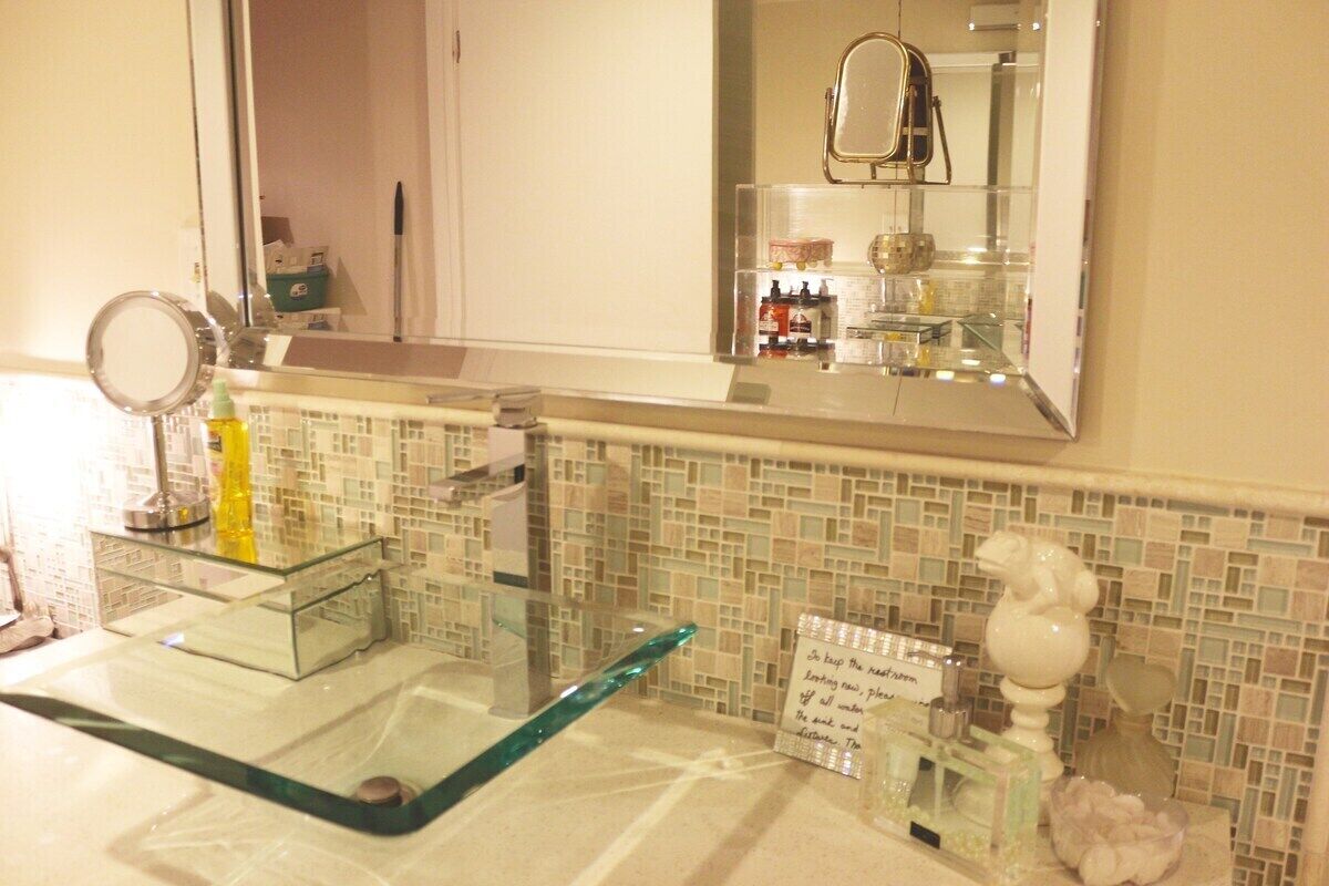 Ein Badezimmer mit einem Waschbecken aus Glas, einem Spiegel, Pflegeartikeln und einer Ziervase.