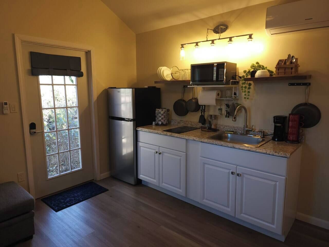 Cocina compacta con armarios blancos, refrigerador de acero inoxidable, microondas y pileta. Hay ollas y sartenes colgando, y una pequeña mesada con cafetera y una taza roja.