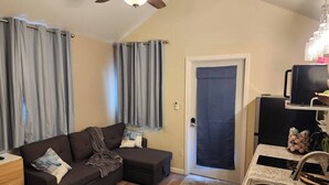 Un espai vital compacte amb un sofà, una cuina petita i una porta que condueix a una altra habitació.