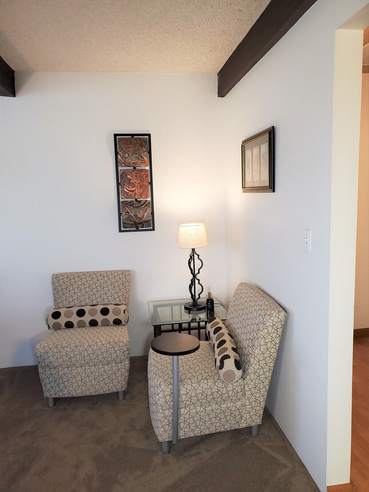 A small living area with two patterned armchairs, a glass-top coffee table, a floor lamp, and two framed artworks on the wall.