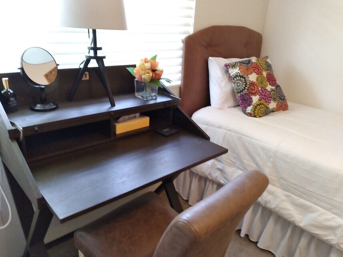 A bedroom with a bed, a desk with a lamp, a chair, and a small table with a vase of flowers.