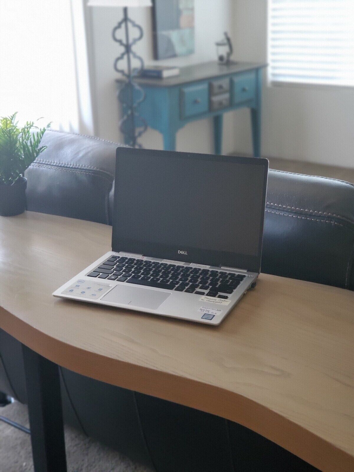 Un ordinateur portable est posé sur une table en bois dans une pièce où l’on aperçoit une chaise bleue, une petite plante et, au fond, un bureau avec des tiroirs.