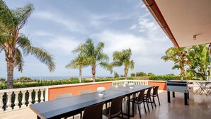 Una terraza con una mesa larga y sillas, con vistas a un paisaje pintoresco con palmeras y un horizonte lejano.