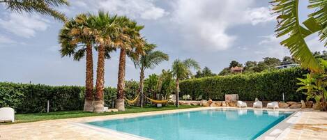 Una piscina rectangular amb una zona de solàrium enrajolat, envoltada de palmeres i un arbustos.