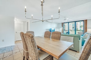 Uma área de refeições moderna com mesa de madeira e cadeiras de vime, ventilador de teto e vista para um edifício residencial através de grandes janelas.