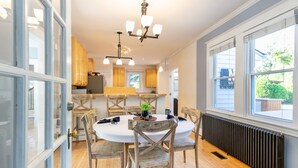 A dining area with a round table and wooden chairs, a chandelier, and a view of the outdoors through large windows. - USNA Home Steps to Stadium Dog-Friendly w Parking (Annapolis)
