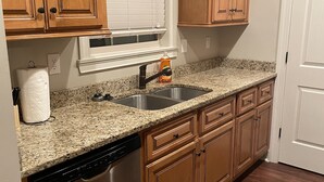 Cocina con gabinetes de madera, lavavajillas de acero inoxidable, encimera de granito y ventana con persianas.