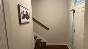 Una escalera de madera con barandilla, que lleva a un rellano con una obra de arte enmarco y una puerta con un ventanal encima.