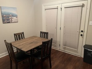Un coin repas avec une table en bois et quatre chaises, un tableau au mur et une porte blanche avec des stores.